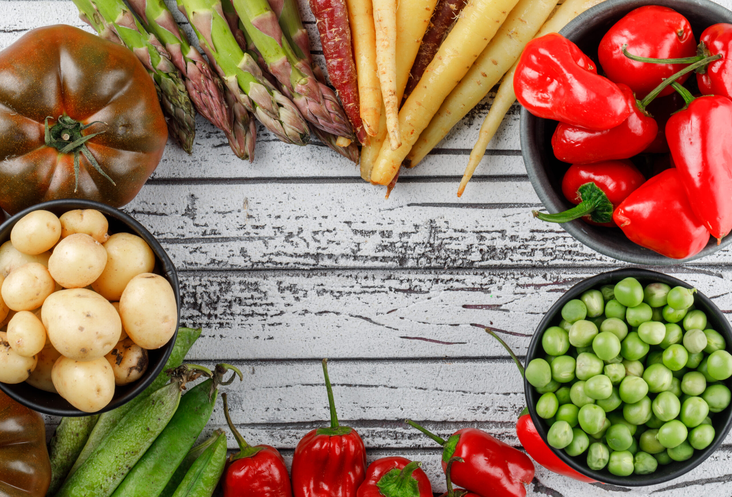 Red peppers with potatoes, tomatoes, asparagus, green pods, peas, carrots in a bowl on wooden background, top view.